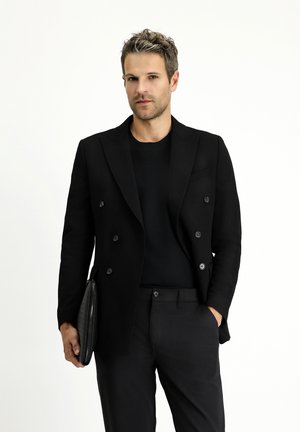 Blazer negro de doble botonadura en una tela suave, combinado con una camiseta negra de cuello redondo y pantalones negros a medida, sosteniendo un clutch texturizado negro.