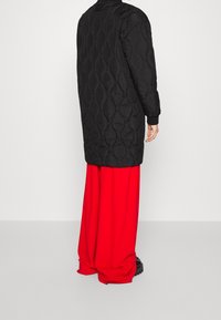 Veste longue matelassée noire avec motif texturé, associée à un pantalon rouge ample et des chaussures à plateforme noires. Caractéristiques : poignets et ourlet côtelés.