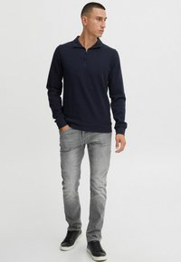 Mornarska dukserica sa zatvaračem i dugim rukavima, u kombinaciji s svijetlosivim trapericama i crnim tenisicama. Outfit ima uski kroj i rebraste manžete.