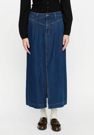 Denim maxi skirt in dark blue. Features front pleats, a button closure, and a center slit. Paired with brown loafers and cream socks.