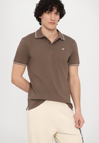 Brown polo shirt with a collar featuring white-striped accents. Small logo on the chest. Worn with light beige pants. Short sleeves.