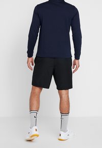 Muž v černých šortkách, tmavě modré dlouhé košili, bílých teniskách a šedých ponožkách Nike stojí na jednoduchém bílém pozadí.