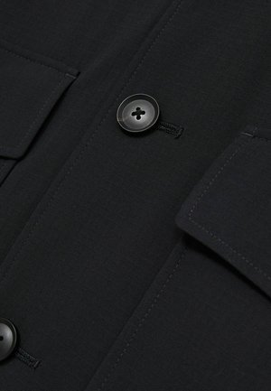 Sort skjortejakke lavet af tekstureret stof, med to lommer og en stor sort knap, samt synlige syninger.