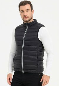 Gilet noir en duvet avec un motif matelassé horizontal, col haut et zip sur le devant. Porté par-dessus une chemise à manches longues blanche. Texture lisse et brillante.