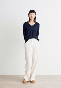 Svetr s V výstřihem v námořnické modři, široké krémové kalhoty a hnědé boty. Měkká látka, volný střih, minimalistický design bez vzorů nebo ozdob.