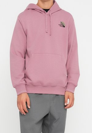 Muž nosící světle fialovou mikinu s kapucí a předním kapsou a malým květinovým logem North Face na hrudi, v kombinaci se šedými kalhotami.