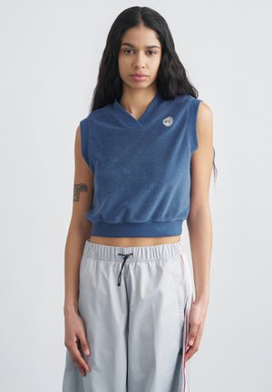 Νεαρή γυναίκα με μακριά σκούρα μαλλιά, φορώντας ένα μπλε αμάνικο κοντό τοπ και ανοιχτό γκρι παντελόνι με κορδόνι, που στέκεται μπροστά σε απλό φόντο.