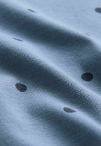 Ljusblått textil med en slät textur, prytt med spridda svarta prickar i varierande storlekar på ytan.