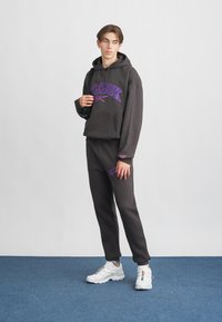 Tumši pelēks džemperis ar purpura krāsas "Reebok" logo, apvienots ar saskaņotām sporta biksēm. Iespējama regulējama josta un diskrēta zīmola norāde uz piedurknēm. Baltas kedas.