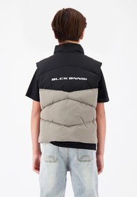 Puffer-Weste in Schwarz und Hellgrau, mit hohem Kragen und gestepptem Muster, hinten mit Logo. Über einem schwarzen Shirt getragen.