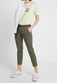T-shirt en coton vert clair avec un imprimé graphique, pantalon fuselé vert olive, veste en denim blanche et baskets blanches avec des accents noirs.