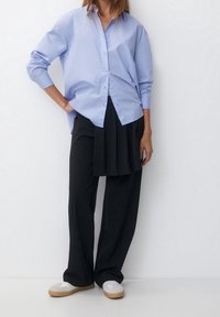 Persona che indossa una camicia azzurro chiaro oversize con bottoni, pantaloni neri a righe con un pannello a gonna attaccato e scarpe da ginnastica bianche.