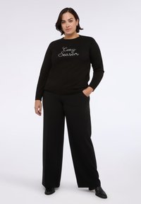 Czarny sweter z białym haftem "Cozy Season", z okrągłym dekoltem i długimi rękawami, w zestawieniu z czarnymi spodniami o szerokich nogawkach i botkami za kostkę.