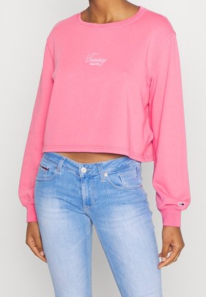 Žena v růžovém zkráceném topu s dlouhým rukávem a logem "Tommy Jeans" a světle modrých džínech s vysokým pasem na jednobarevném pozadí.