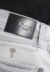Detailní záběr na kapsu světle šedých džínových kalhot s černou koženou značkou Pepe Jeans London a kovovým knoflíkem.