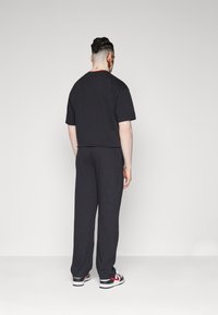 Uomo con capelli intrecciati che indossa una maglietta nera, pantaloni neri e scarpe bianche con swoosh rosso, in piedi di fronte a un muro bianco semplice.