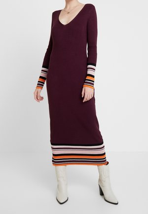 Longue robe en maille côtelée bordeaux avec col en V, poignets et ourlet rayés noir, rose, orange, portée avec des bottes blanches à talons.