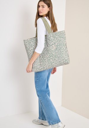 Mujer con camiseta blanca y vaqueros azules lleva un gran bolso tote gris con estampado de leopardo verde y asas verde oliva.