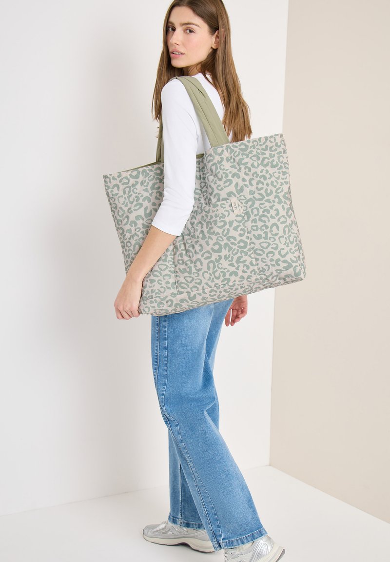 Femme portant un haut blanc et un jean bleu, portant un grand sac cabas gris avec un imprimé léopard vert et des anses vert olive.
