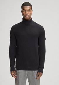 Pull en maille à col roulé de couleur bleu marine foncé, avec un tissu texturé, des poignets et un ourlet côtelés. Étiquette de marque discrète sur la manche gauche.