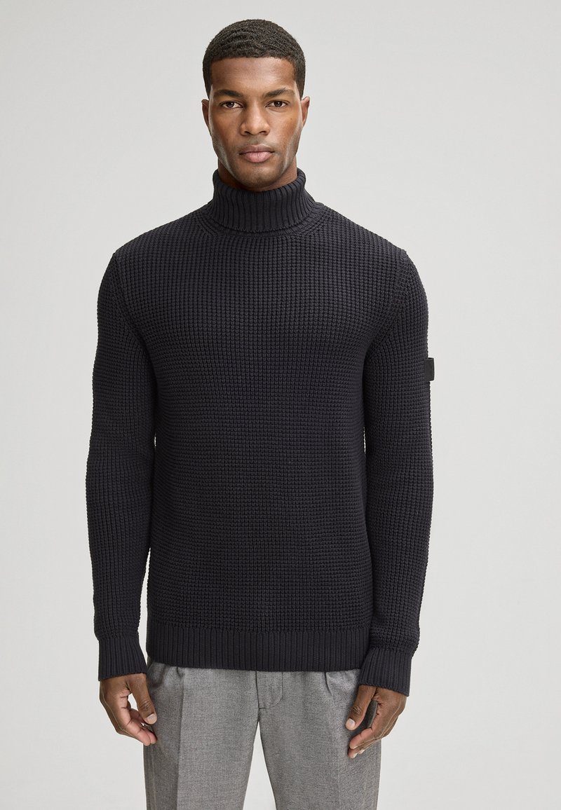 Pull en maille à col roulé de couleur bleu marine foncé, avec un tissu texturé, des poignets et un ourlet côtelés. Étiquette de marque discrète sur la manche gauche.