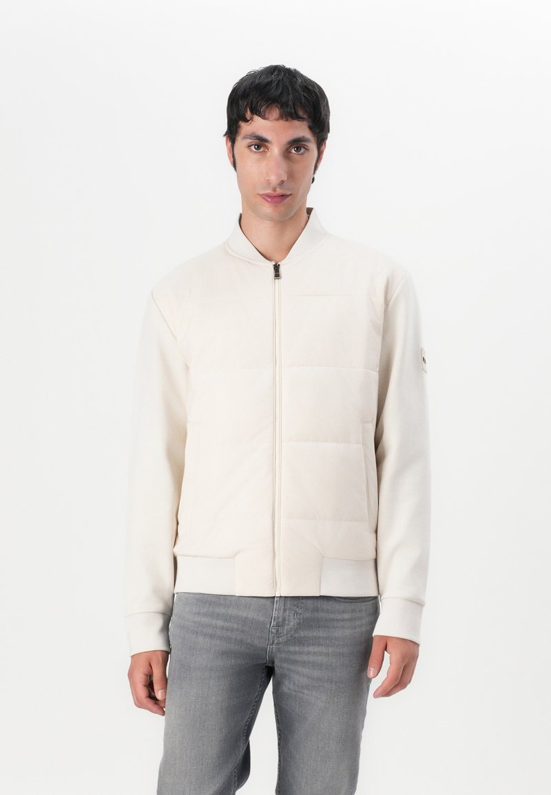 Hellbeige Bomberjacke mit Reißverschluss vorne, gepolstertem Steppbereich, gerippten Bündchen und zwei seitlichen Taschen. Kombiniert mit grauen Jeans.