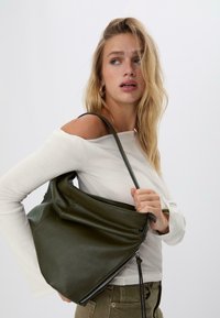 Borsa a tracolla in pelle verde con una texture morbida, forma strutturata e dettaglio con zip. Modello che indossa una maglietta bianca a spalle scoperte e pantaloni oliva.
