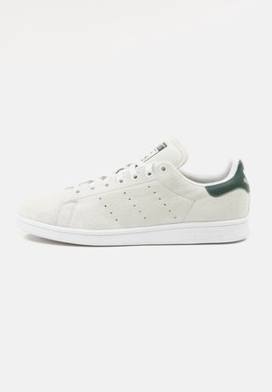 Biały sneaker Adidas Stan Smith z zielonym paskiem na pięcie, perforowanymi bocznymi paskami, białą podeszwą i sznurowaniem, pokazany z boku na białym tle.