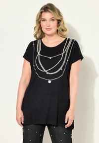 Schwarzes T-Shirt mit kurzen Ärmeln, ausgestattet mit einem Kettenmuster in Silber und Creme. Weicher Stoff mit lockerem Schnitt und rundem Ausschnitt.