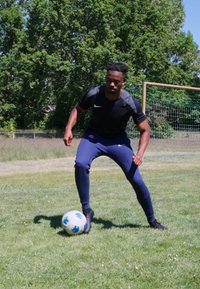 Atleta con una camiseta negra de Nike y pantalones deportivos azules, mostrando una postura dinámica mientras controla un balón de fútbol blanco y azul sobre la hierba.