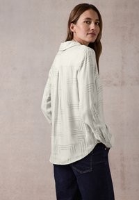Maglietta a maniche lunghe grigia chiara con un motivo geometrico, colletto e texture morbida, abbinata a pantaloni in denim scuri.