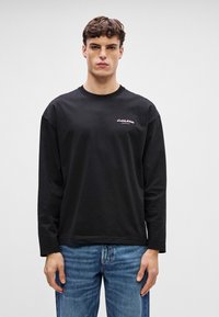 Svart langermet T-skjorte i bomull, med rund hals og en liten rosa logo på brystet. Kombinert med blå jeans.