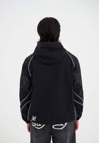 Giacca nera con cappuccio, caratterizzata da un design testurizzato sulle maniche, dettagli bianchi e un logo sulla parte bassa della schiena. Il tessuto sembra leggero.