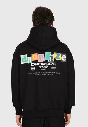 Person in einem schwarzen Hoodie mit bunten "dropsize" Buchstaben und weißem Text auf dem Rücken, der "DROPSIZE DESIGNS EST. 2020" liest.