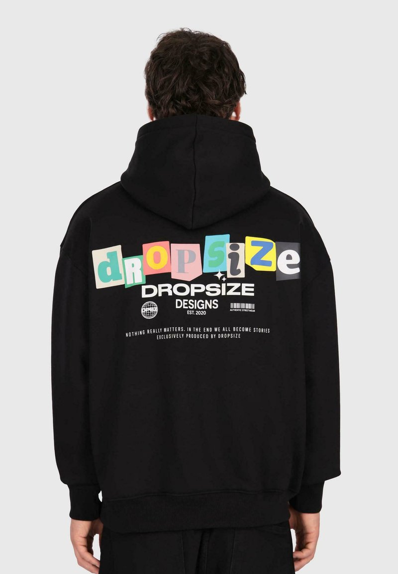 Personne portant un sweat à capuche noir avec des lettres colorées "dropsize" et du texte blanc indiquant "DROPSIZE DESIGNS EST. 2020" dans le dos.