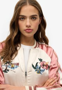 Kurtka bomber z różowymi satynowymi rękawami, kremowym tułowiem i haftem kwiatowym. Posiada zamek błyskawiczny z przodu oraz prążkowany kołnierz w paski.