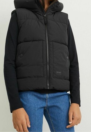 Veste matelassée noire avec fermeture éclair à l'avant, capuche et poches à boutons-pression. Portée sur un haut noir à manches longues et un jean bleu. Tissu texturé.