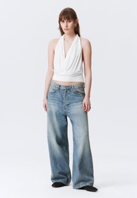 Weißes Top mit Halterneck und Drapé-Design, kombiniert mit lockeren, hellblauen Jeans aus Denim mit einer ausgewaschenen Optik und weitem Bein. Schwarze Schuhe.