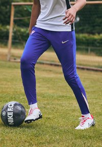 Violet sportiniai kelnės su baltais ir mėlynais akcentais, derinami su baltais futbolo batais su raudonais raišteliais, šalia juodo futbolo kamuolio.