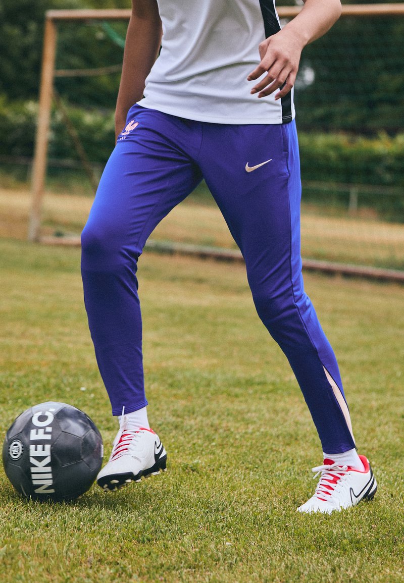 Violet sportiniai kelnės su baltais ir mėlynais akcentais, derinami su baltais futbolo batais su raudonais raišteliais, šalia juodo futbolo kamuolio.