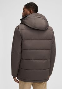 Giacca a vento in marrone scuro, design trapuntato, cappuccio grande e texture liscia. Presenta un dettaglio con logo sulla manica sinistra.