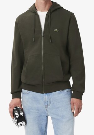 Homme portant un sweat à capuche zippé vert foncé avec un logo crocodile, jean bleu clair, tenant une petite pochette noire et blanche dans la main gauche.