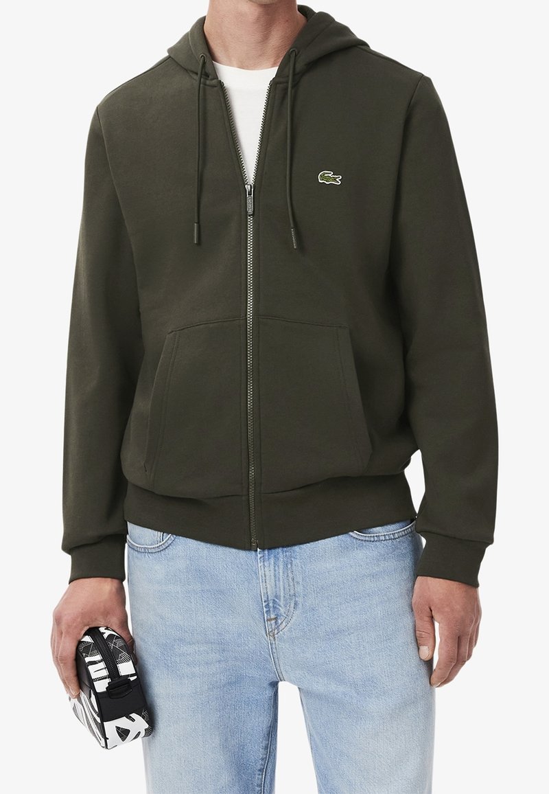 Homme portant un sweat à capuche zippé vert foncé avec un logo crocodile, jean bleu clair, tenant une petite pochette noire et blanche dans la main gauche.