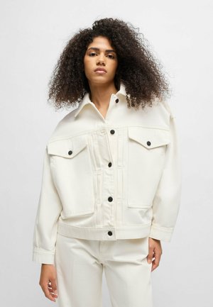 White denim jacket with a high collar, large pockets, and black buttons. Smooth texture; loose fit; paired with matching white pants.
