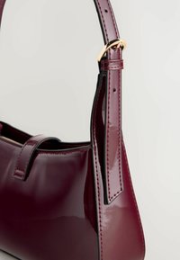 Bordeaux patenteret læderhandbag med en struktureret design, der har et fladt håndtag, guld hardware detaljer og kontrasterende sort syning.