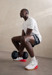 Sort sekskantet håndvægt holdt af en person i en hvid skjorte og sorte shorts med hvid skrift, iført hvide sko med røde detaljer.
