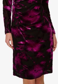 Detailní záběr na černé šaty s velkými, živými fialovými květinovými vzory, ukazující dlouhé rukávy a dolní část trupu až po střed stehna.