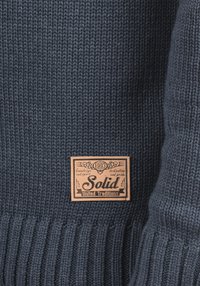 Marineblauer Strickpullover mit strukturiertem Muster und einem aufgenähten Lederetikett mit der Aufschrift "Solid United Traditions 1993".