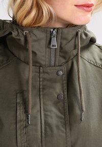 Olijfgroene jas met rits aan de voorkant en capuchon. Voorzien van metalen hardware, verstelbare trekkoorden en drukknopaccenten. Getextureerde stof.