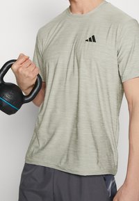 Ljusgrå idrottströja med en slät textur, prydd med en liten svart logotyp på bröstet, i kombination med en svart kettlebell i handen.